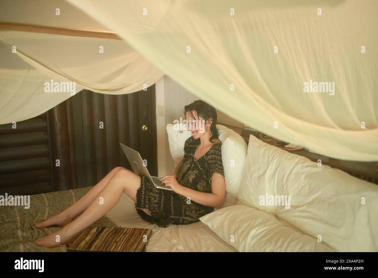 Une femme est assise sur un lit avec des draps blancs, engagée dans l'utilisation de son ordinateur portable. Elle est sous une moustiquaire douce et transparente, portant des lunettes et des vêtements décontractés, avec une posture détendue dans une pièce confortable et faiblement éclairée. Banque D'Images