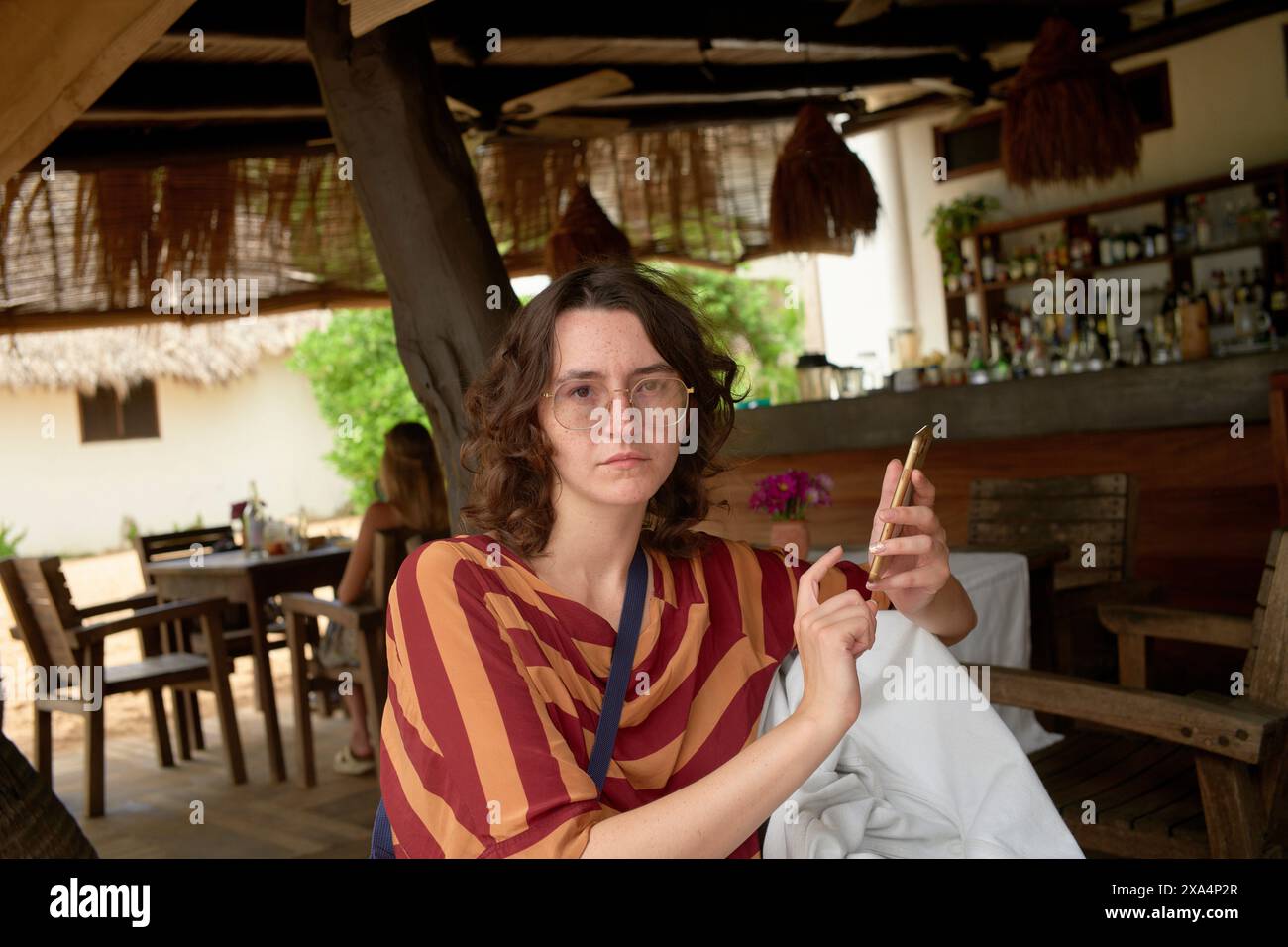 Une femme est assise dans un bar rustique tenant une cigarette ; derrière elle se trouve une étagère en bois avec des bouteilles assorties et un toit de chaume, donnant à la scène une atmosphère décontractée et tropicale. Banque D'Images