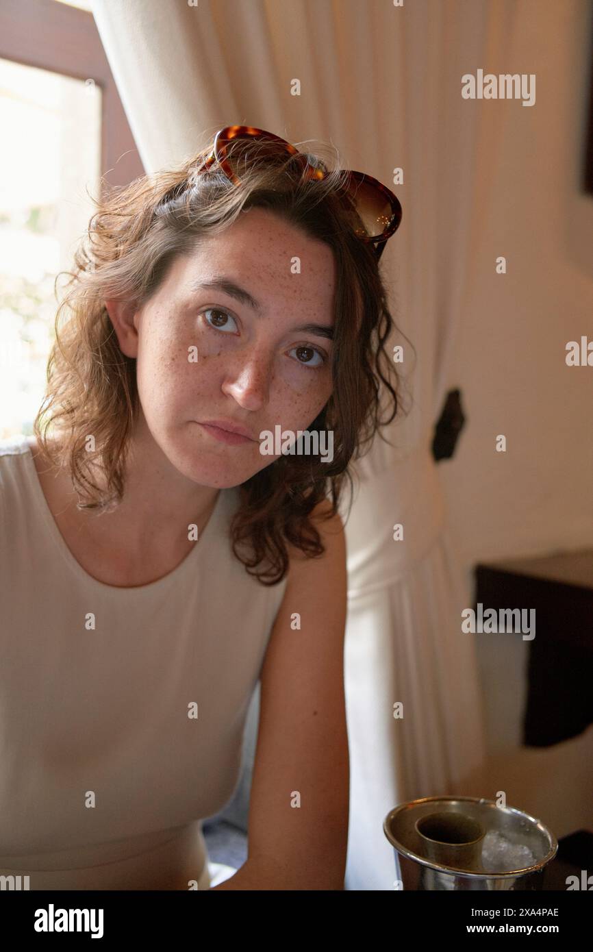 Une femme avec des cheveux bouclés et des lunettes de soleil sur la tête est assise à l'intérieur, avec un léger sourire et une expression contemplative. Banque D'Images