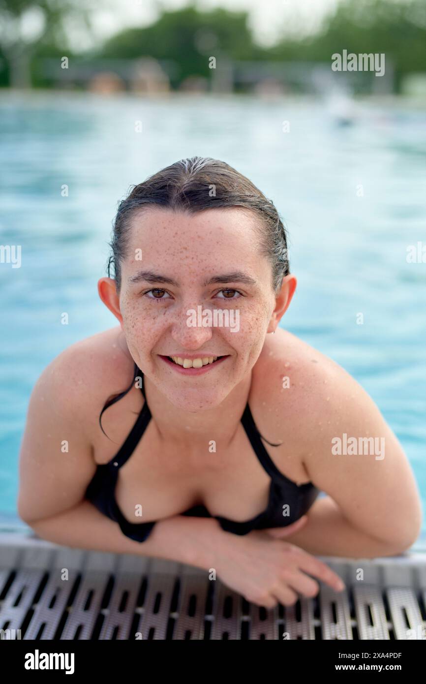 Une jeune femme souriante aux cheveux mouillés est dans une piscine, se reposant sur le bord de la piscine et regardant directement la caméra. Banque D'Images