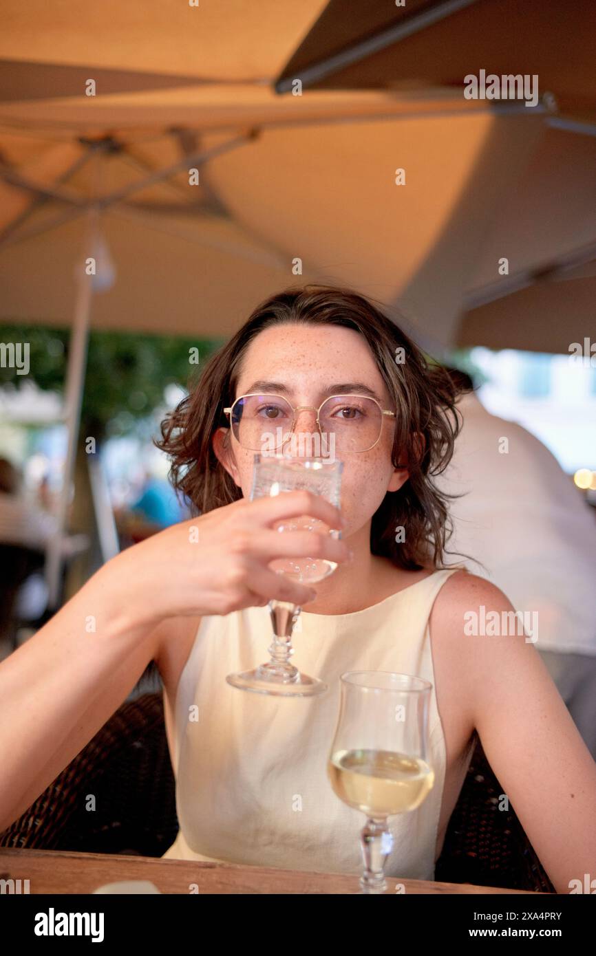 Une personne est assise à une table sous un parapluie, tenant un verre de vin près de son visage. Banque D'Images