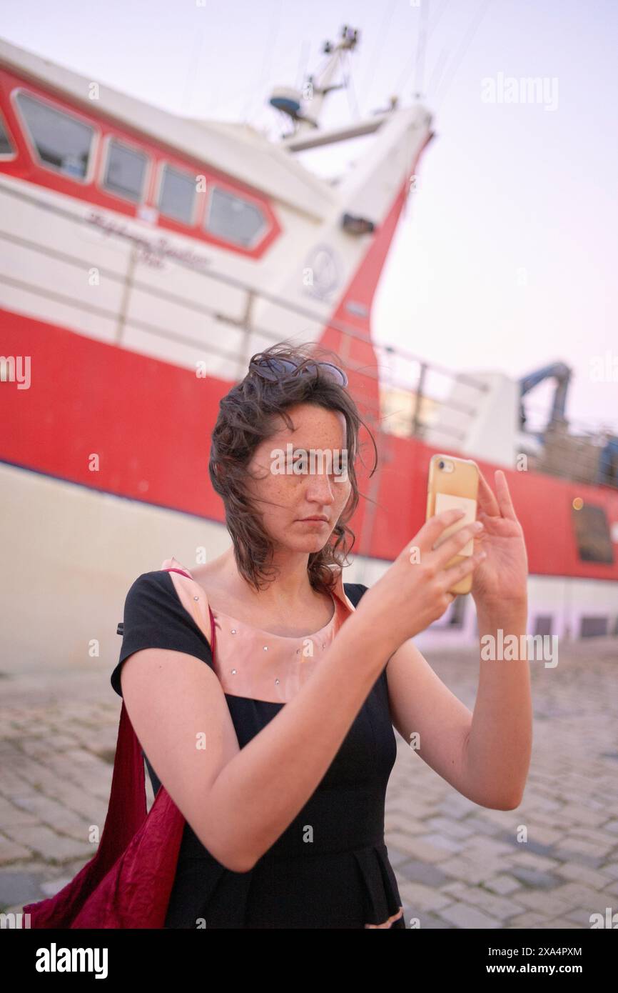 Une femme prend un selfie avec un smartphone devant un bateau rouge et blanc. Banque D'Images