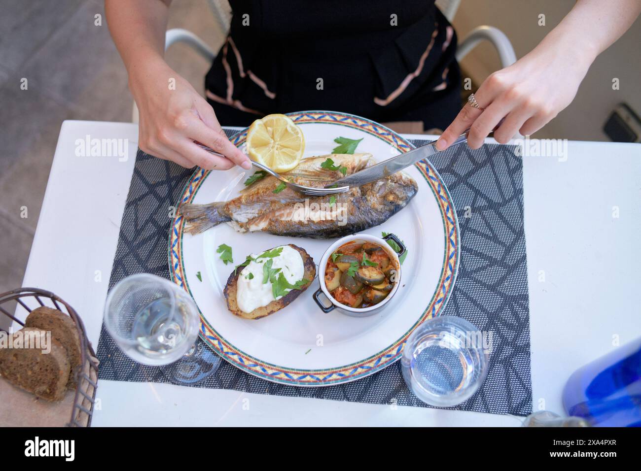 Une personne est assise à une table, sur le point de profiter d'un repas composé d'un poisson grillé garni de citron, d'un côté de pain, d'une sauce crémeuse, et d'un petit bol de ce qui ressemble à des légumes cuits. Banque D'Images