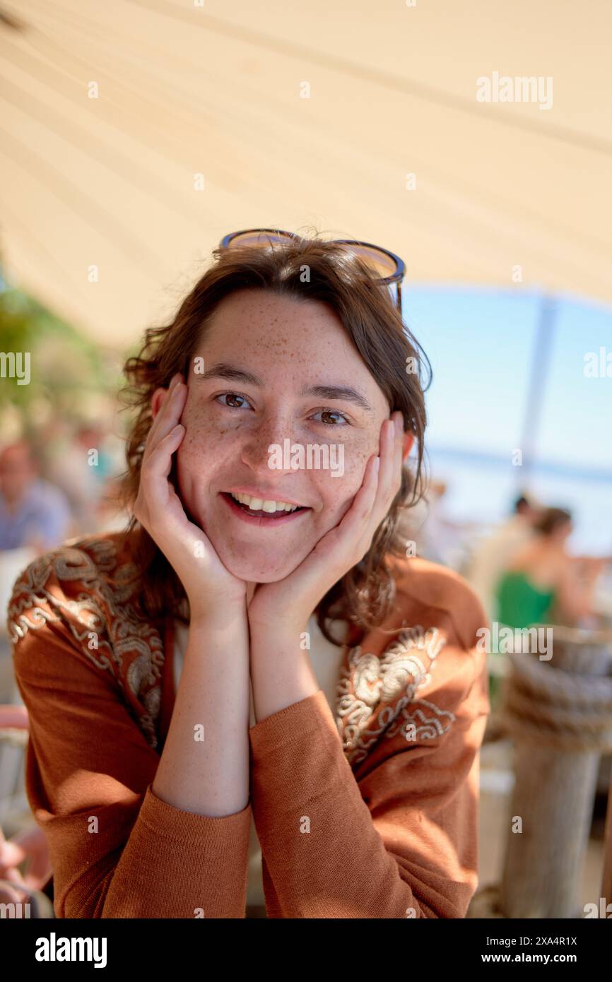 Une femme sourit à la caméra, posant son menton sur ses mains, avec un café extérieur ensoleillé en arrière-plan. Banque D'Images