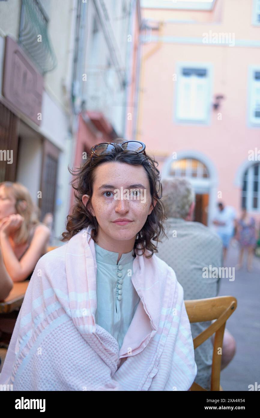 Une femme aux cheveux bouclés portant des lunettes de soleil sur la tête et un dessus bleu clair est assise dans un café en plein air, avec un bâtiment rose en arrière-plan. Banque D'Images