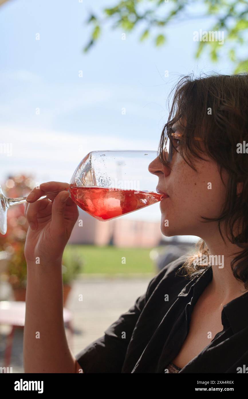 Une jeune femme sirotant du vin rose dans un verre, assise à l'extérieur avec des bâtiments flous en arrière-plan. Banque D'Images