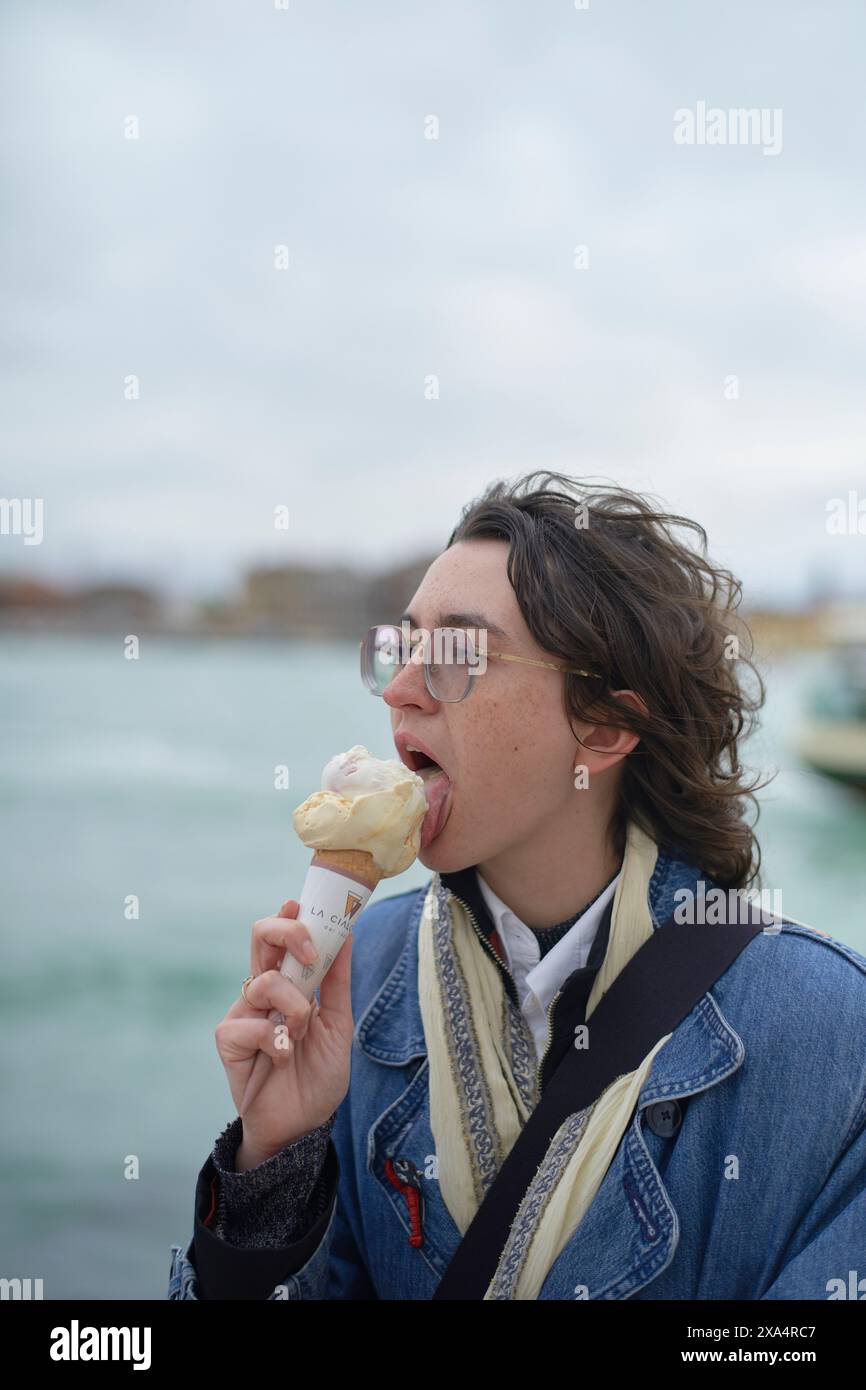 Jeune adulte profitant d'un cône de crème glacée au bord de l'eau avec des bateaux en arrière-plan. Banque D'Images