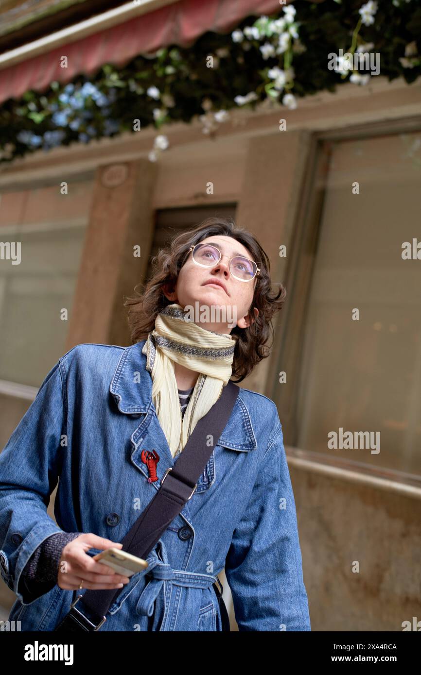 Femme curieuse avec des lunettes regardant vers le haut tenant un smartphone dans une rue de la ville. Banque D'Images