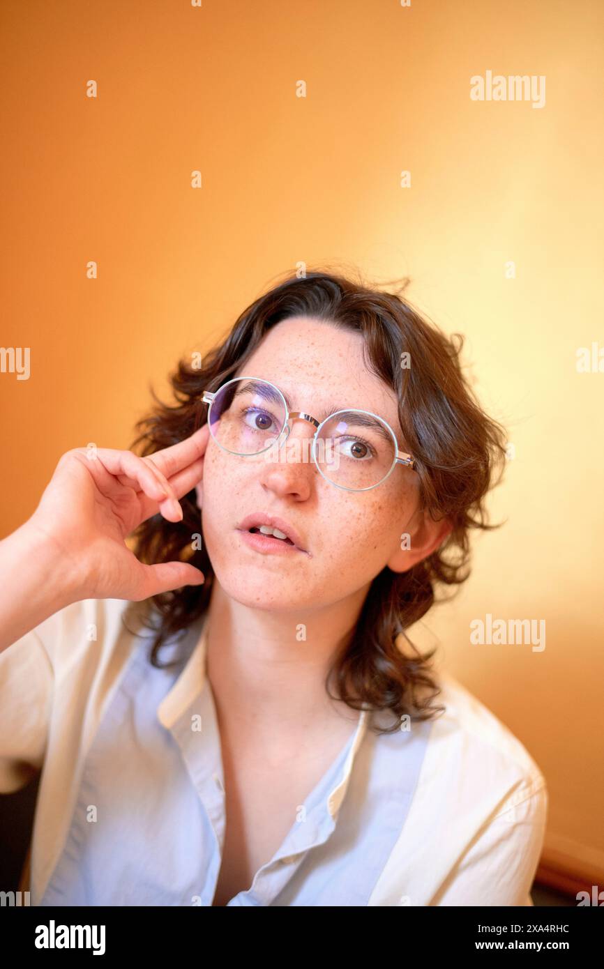 Femme curieuse avec des lunettes qui font des gestes tout en regardant vers le haut sur un fond aux tons chauds. Banque D'Images