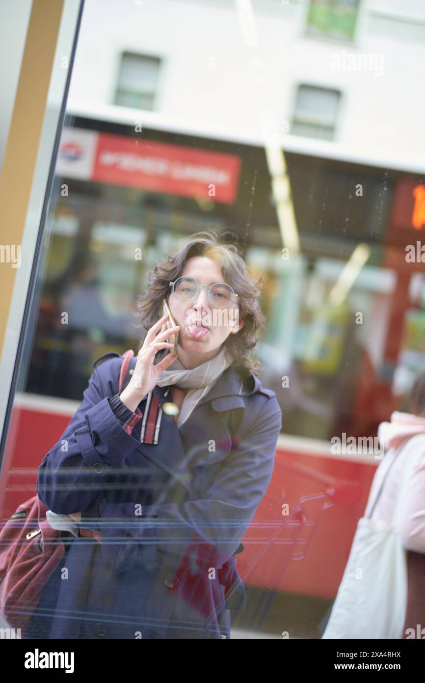 Femme espiègle sortant sa langue lors d'un appel téléphonique à travers un reflet de verre. Banque D'Images