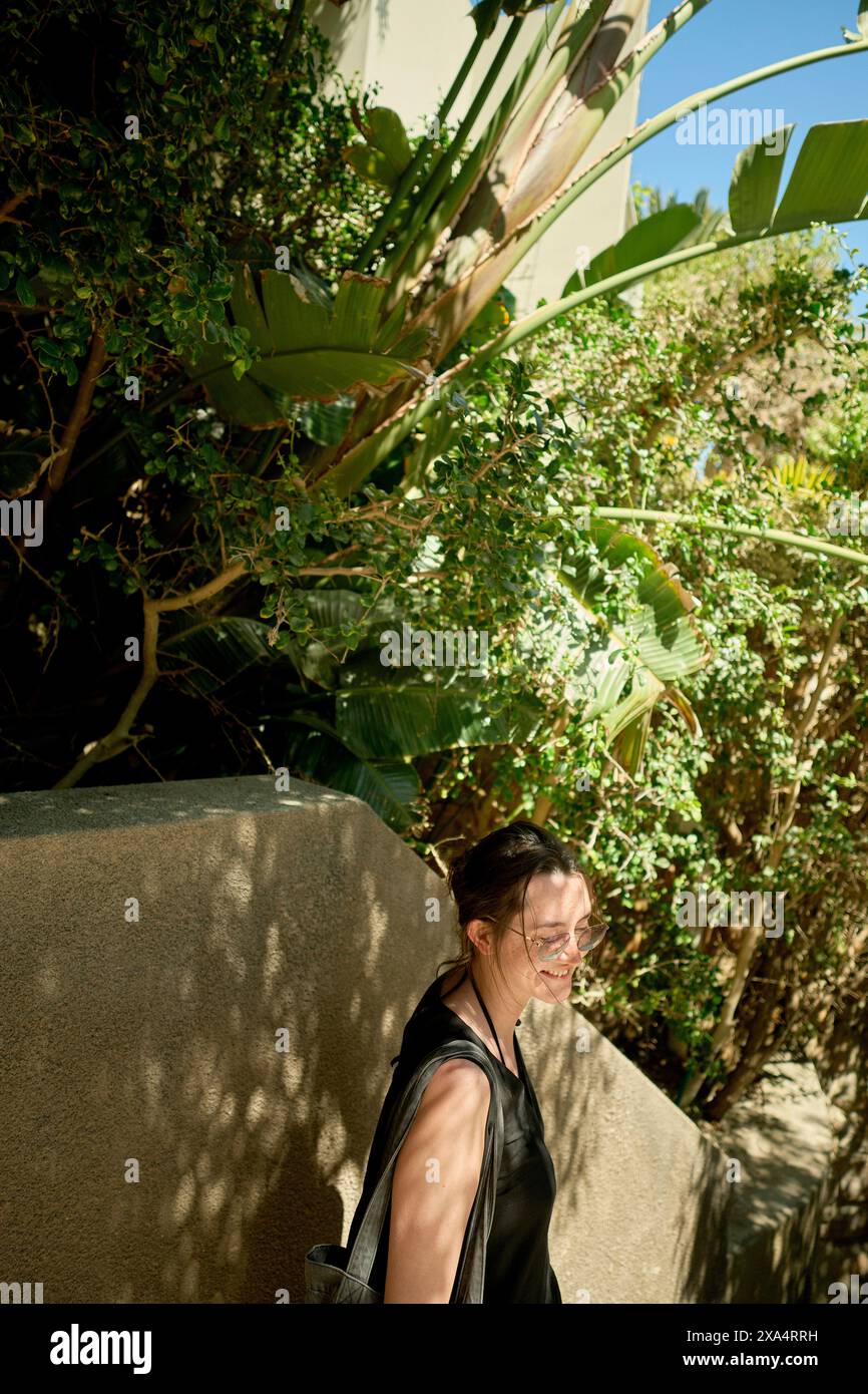 Une femme souriante marche près d'un mur avec des plantes vertes luxuriantes en arrière-plan par une journée ensoleillée. Banque D'Images