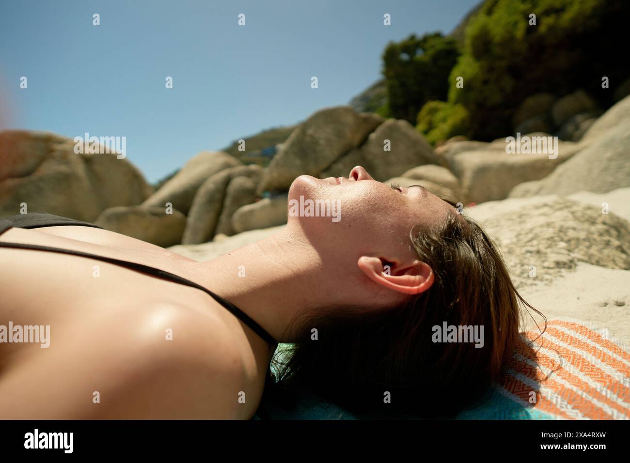 Femme se relaxant sur une serviette de plage dans un contexte de verdure luxuriante et de rochers ensoleillés. Banque D'Images