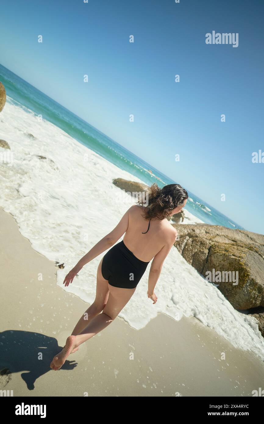 Une femme en maillot de bain noir marche vers la mer sur une plage ensoleillée avec des vagues qui s'écrasent sur les rochers. Banque D'Images