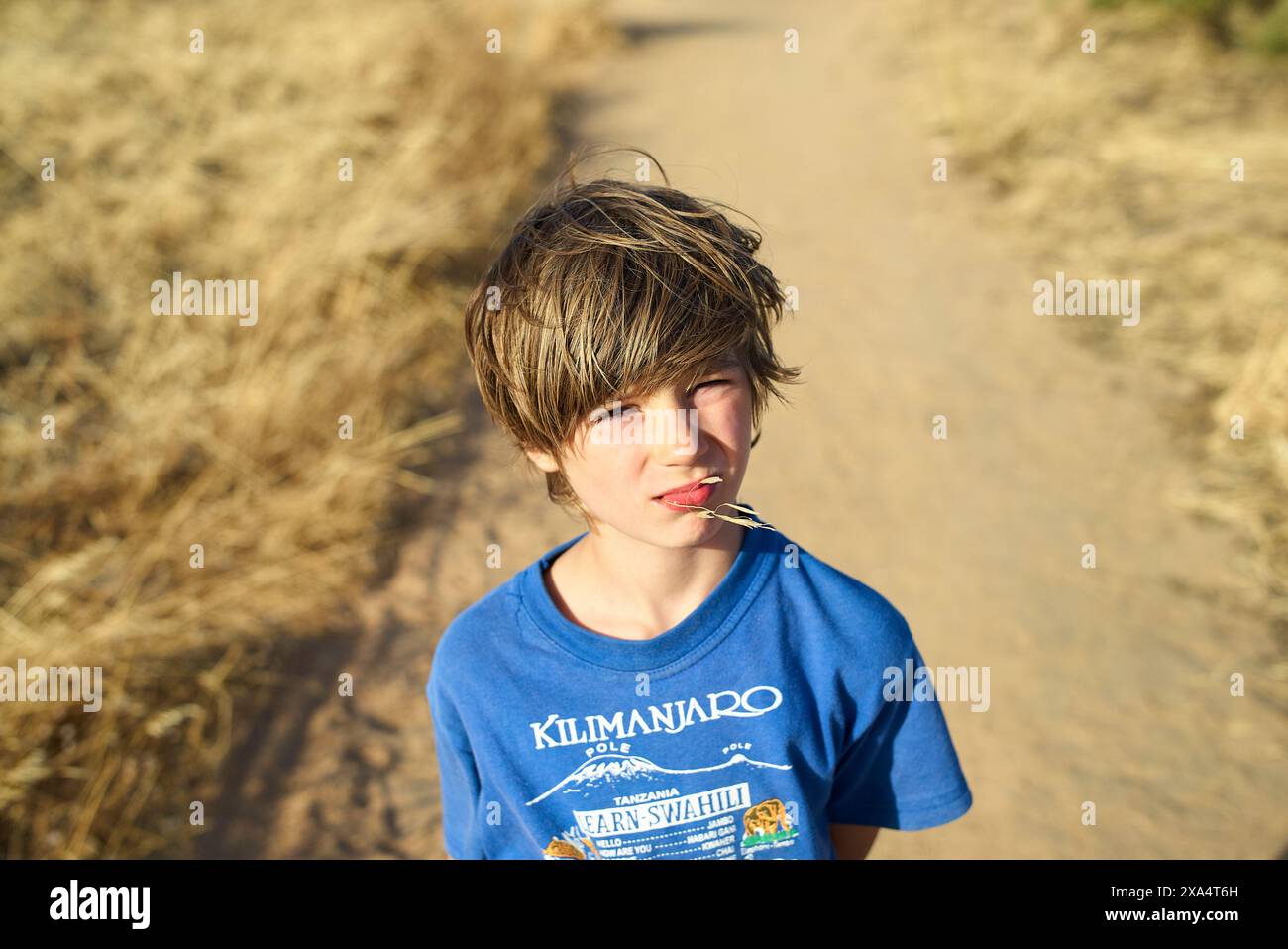 Jeune garçon avec des cheveux touffus debout sur une route poussiéreuse mâchant une paille, avec de l'herbe sèche de chaque côté. Banque D'Images