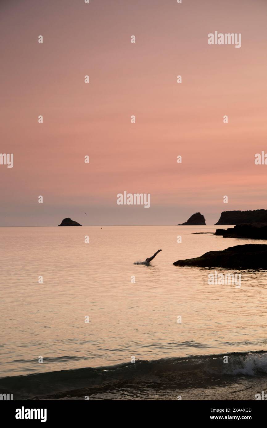 Les teintes crépusculaires peignent le ciel et l'océan dans des tons de rose et d'orange tandis que des formations rocheuses ponctuent la mer tranquille. Banque D'Images