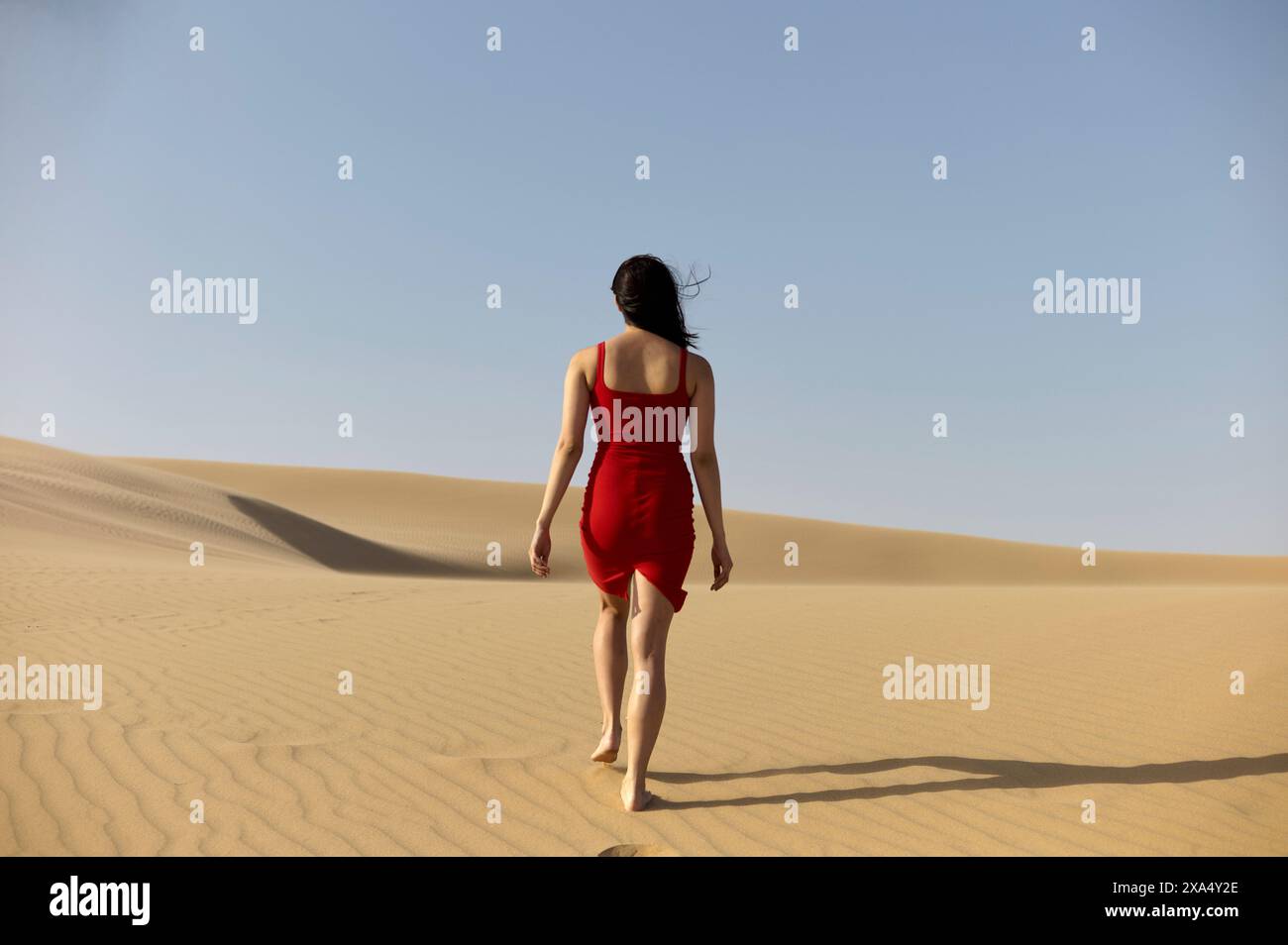 Femme dans une robe rouge marchant sur une dune de sable sous un ciel clair. Banque D'Images