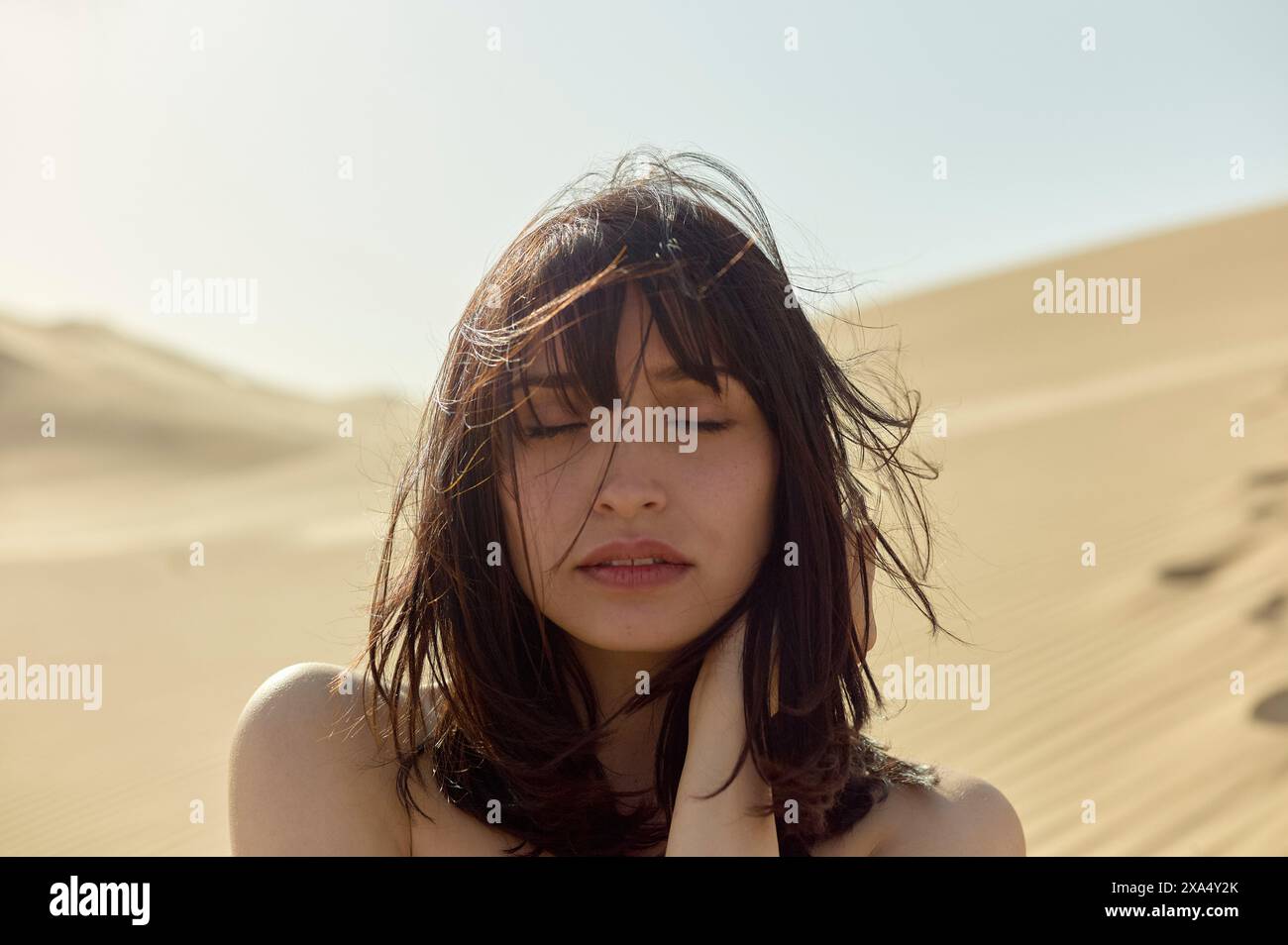 Femme embrassée par le soleil avec les yeux fermés et les cheveux tourbillonnés par le vent se tient au milieu du sable doré du désert. Banque D'Images