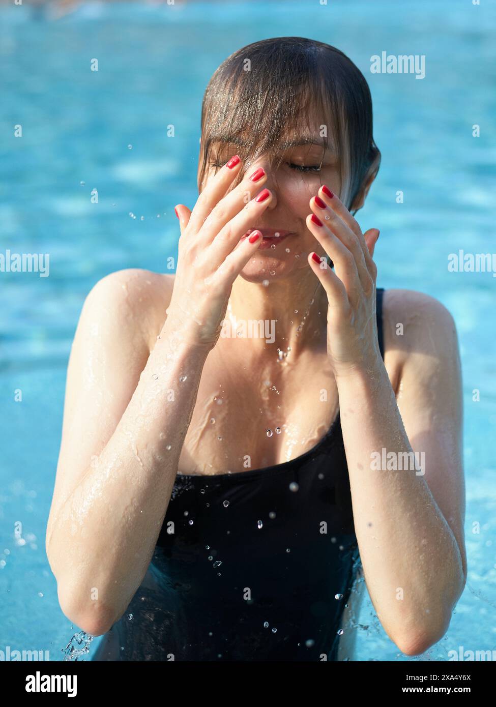 Une femme émerge d'une piscine, essuyant l'eau de son visage avec ses mains, ses cheveux mouillés accrochés à son front. Banque D'Images