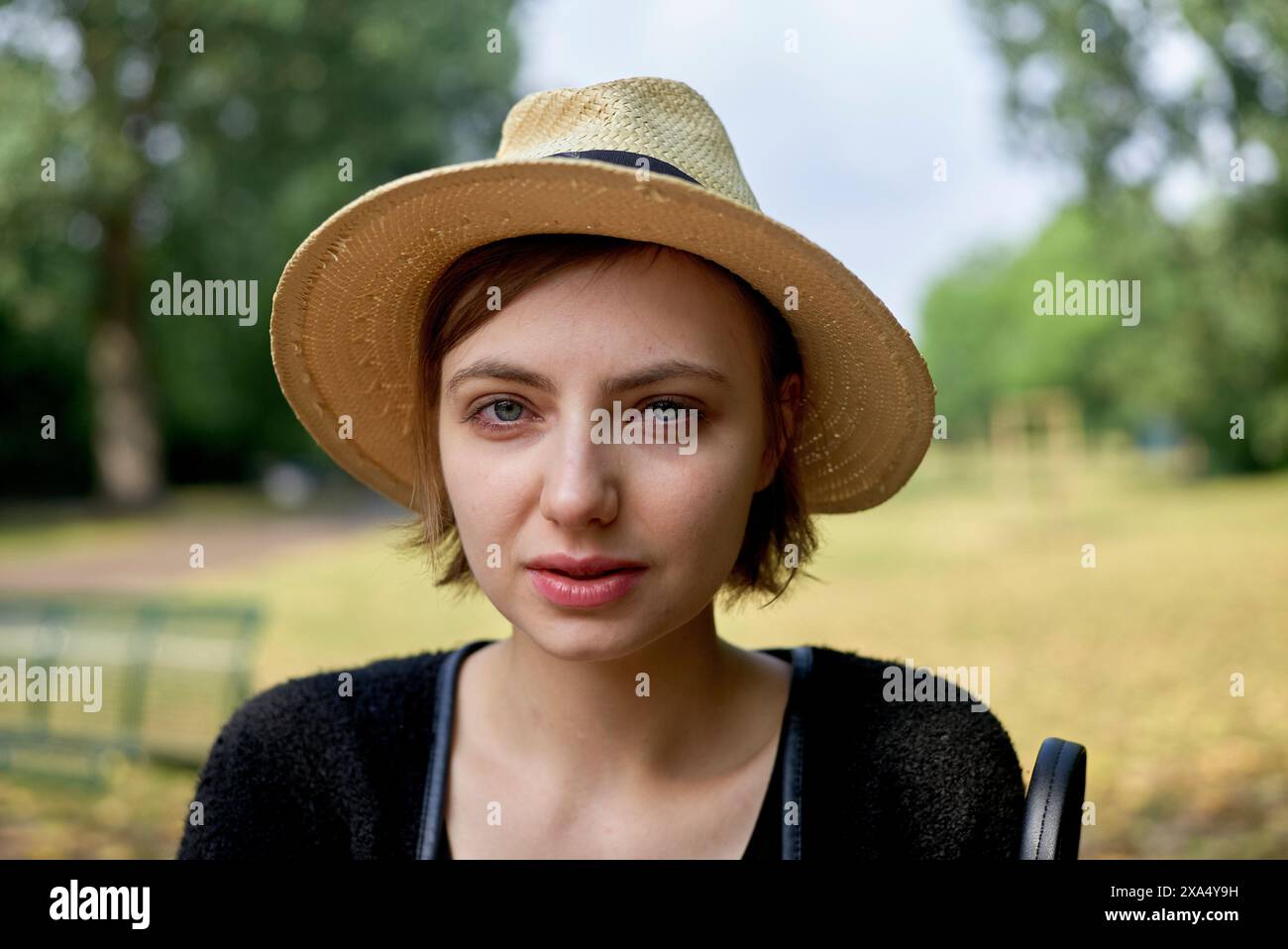 Femme avec un chapeau de paille et top noir souriant dans un parc avec des arbres verts en arrière-plan. Banque D'Images