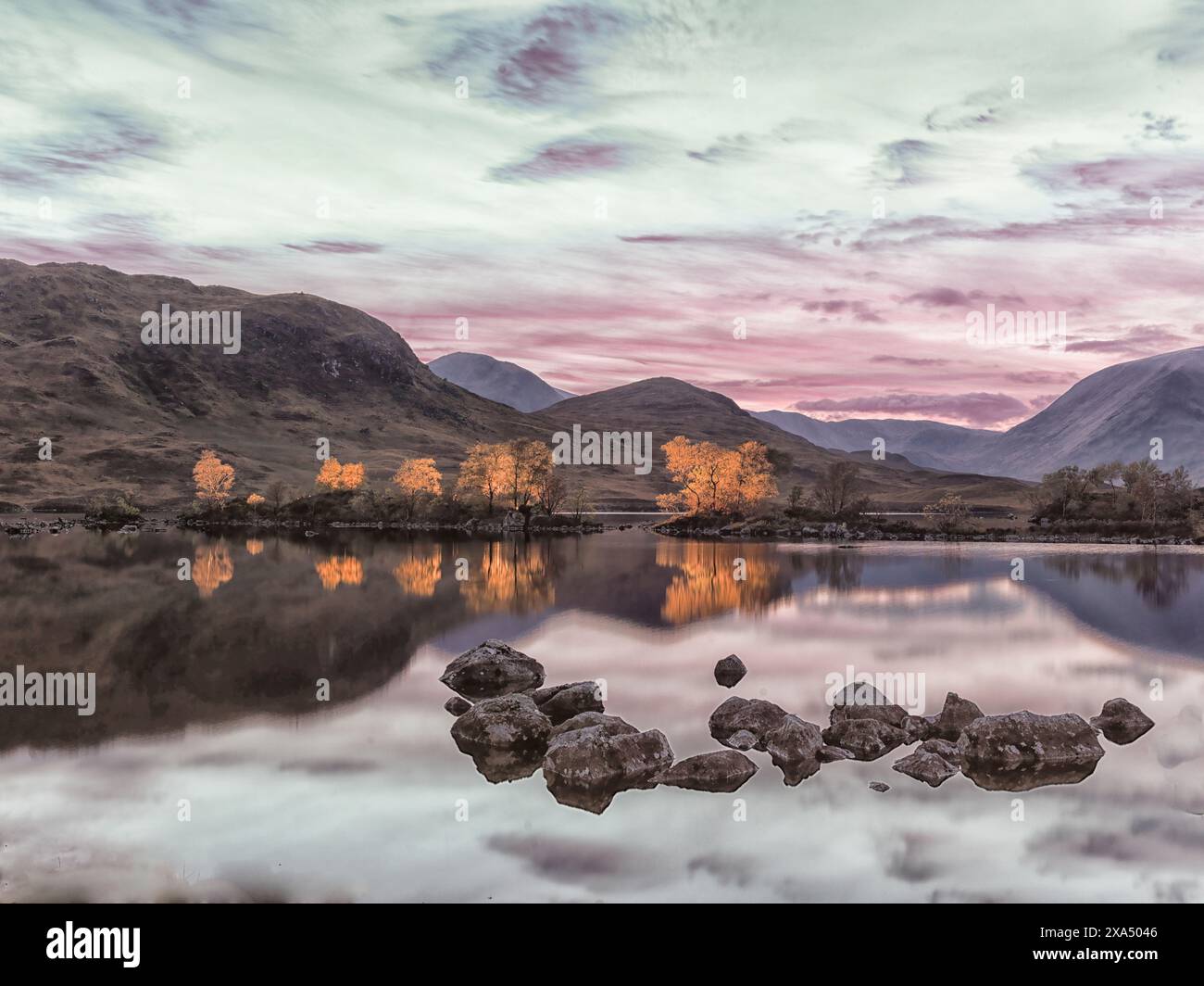 Coucher de soleil tranquille sur un lac en miroir avec des rochers au premier plan et des montagnes en arrière-plan sous un ciel coloré. Banque D'Images