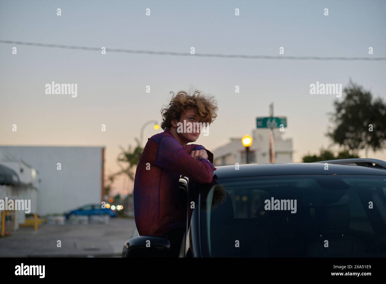 Jeune personne avec les cheveux bouclés appuyé sur le toit d'une voiture au crépuscule dans un cadre urbain. Banque D'Images