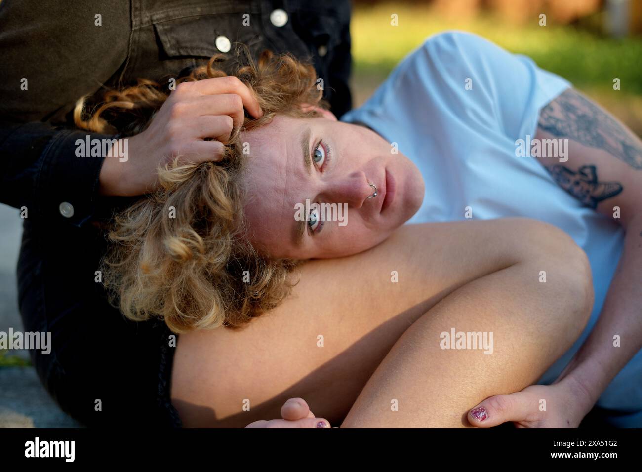 Personne aux cheveux bouclés avec un anneau nasal et un tatouage s'incline sur l'herbe, regardant vers le haut réfléchi. Banque D'Images