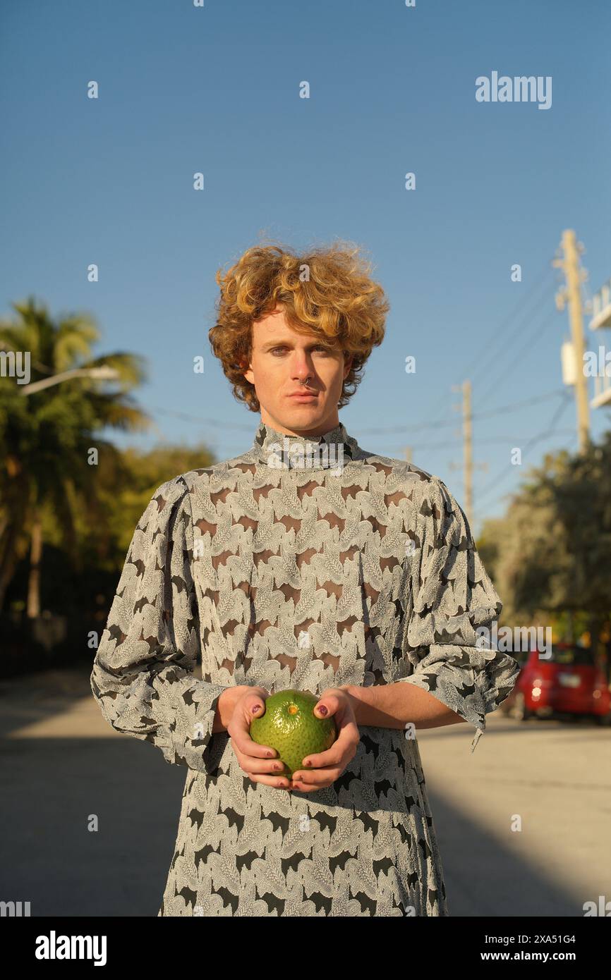 Jeune homme aux cheveux bouclés debout à l'extérieur tenant une pomme verte, portant une chemise à motifs avec des arbres. Banque D'Images