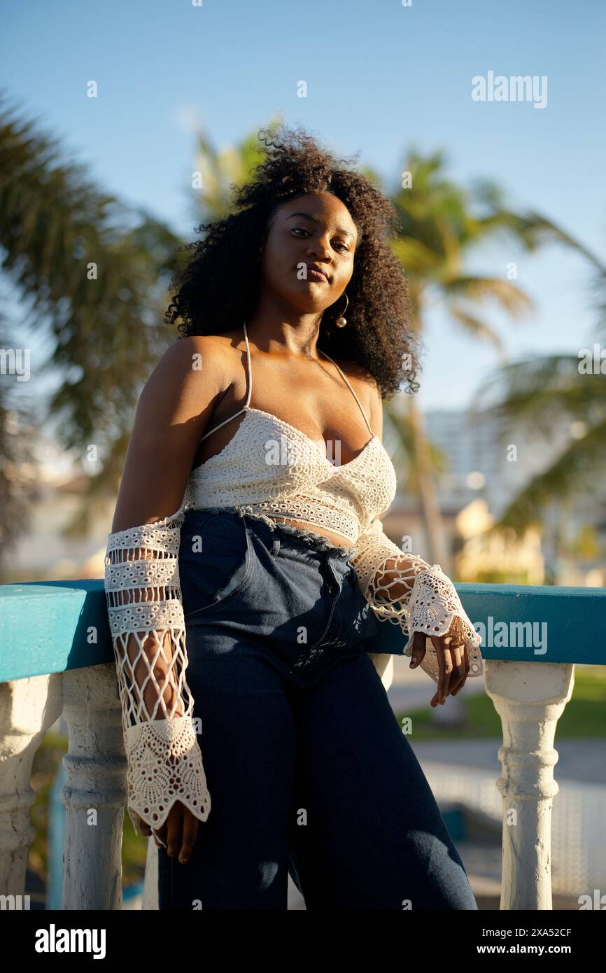 Une femme confiante s'appuie contre une balustrade avec un fond tropical, portant un haut en crochet blanc et un denim. Banque D'Images