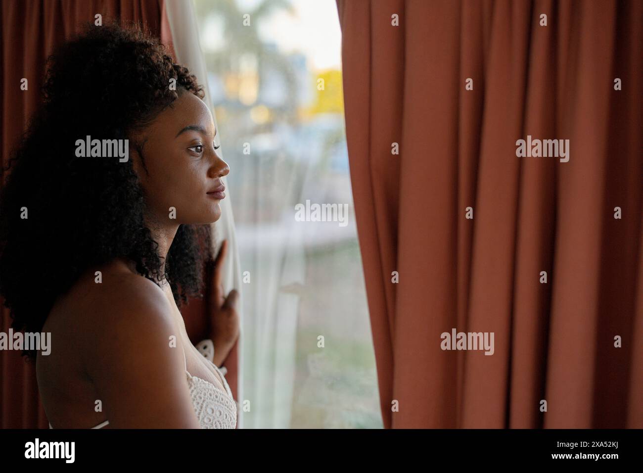 Une femme réfléchie regardant par une fenêtre avec des rideaux transparents partiellement dessinés, avec une lumière naturelle douce soulignant son profil. Banque D'Images