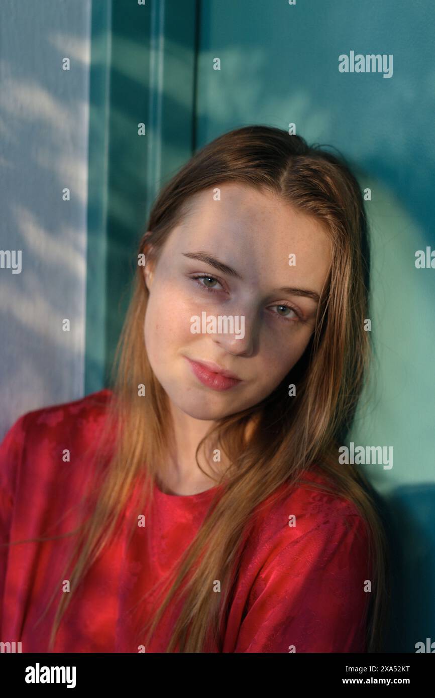 Jeune femme aux longs cheveux blonds portant une chemise rouge, souriant doucement à la lumière naturelle avec un motif d'ombre sur son visage. Banque D'Images