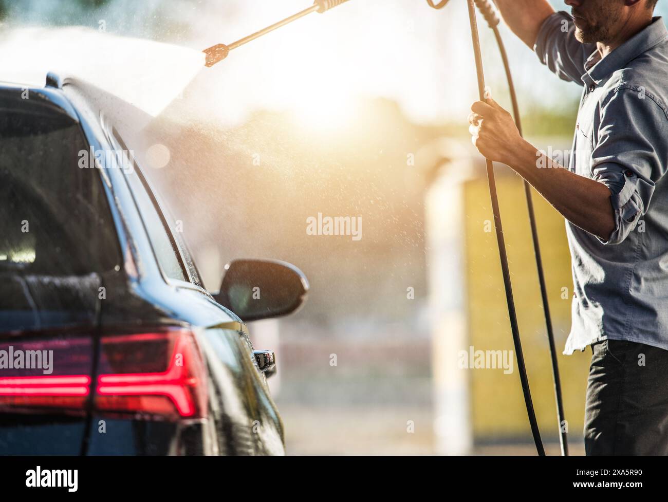 Un ouvrier de lavage de voiture caucasien nettoyant l'extérieur de la voiture des clients en pulvérisant de l'eau et en rinçant la saleté Banque D'Images