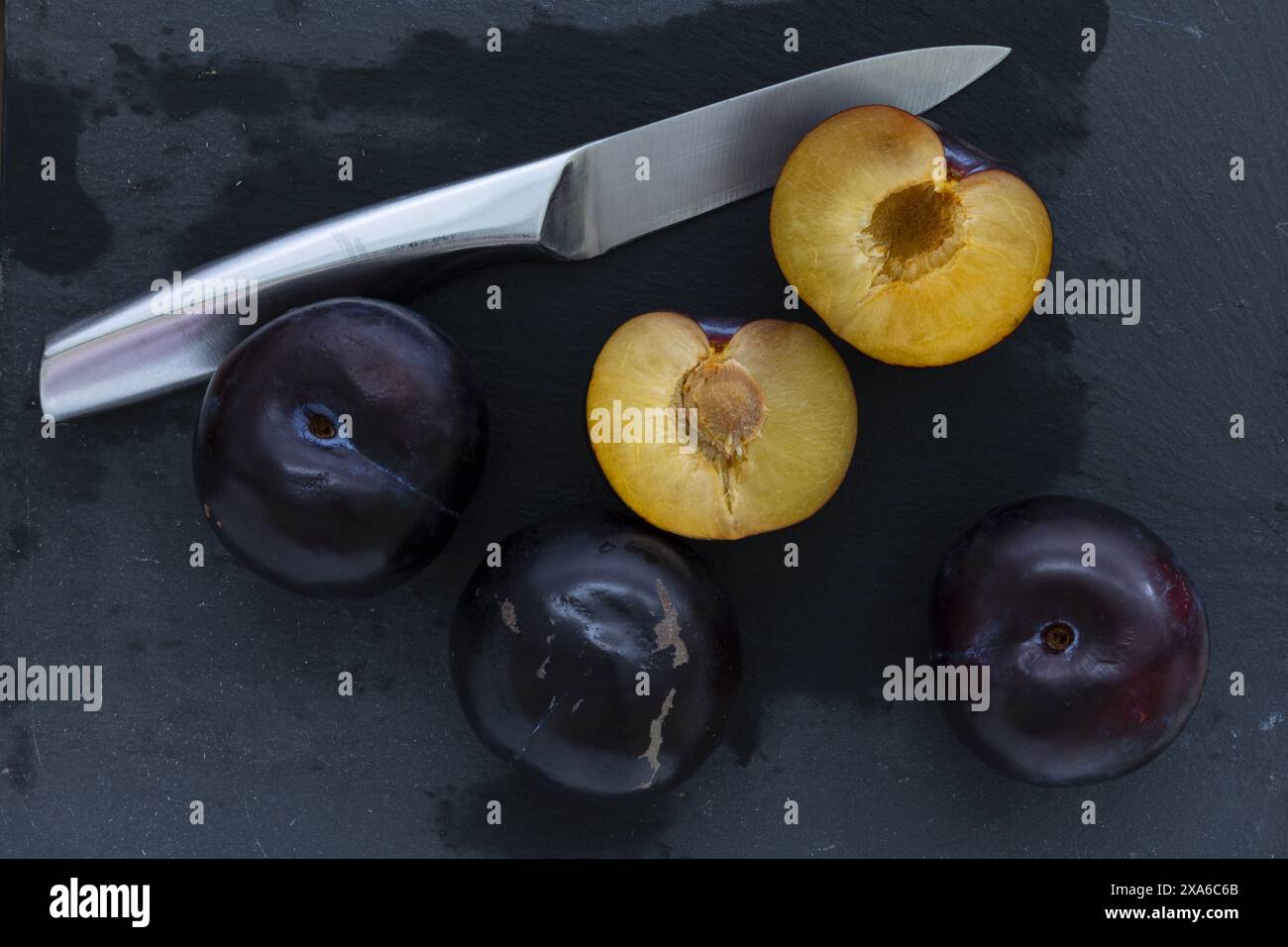 photo de table en gros plan de demi-fruits entiers et coupés de prune bleu foncé avec couteau en acier sur un fond sombre, photographie de natures mortes alimentaires discrètes Banque D'Images