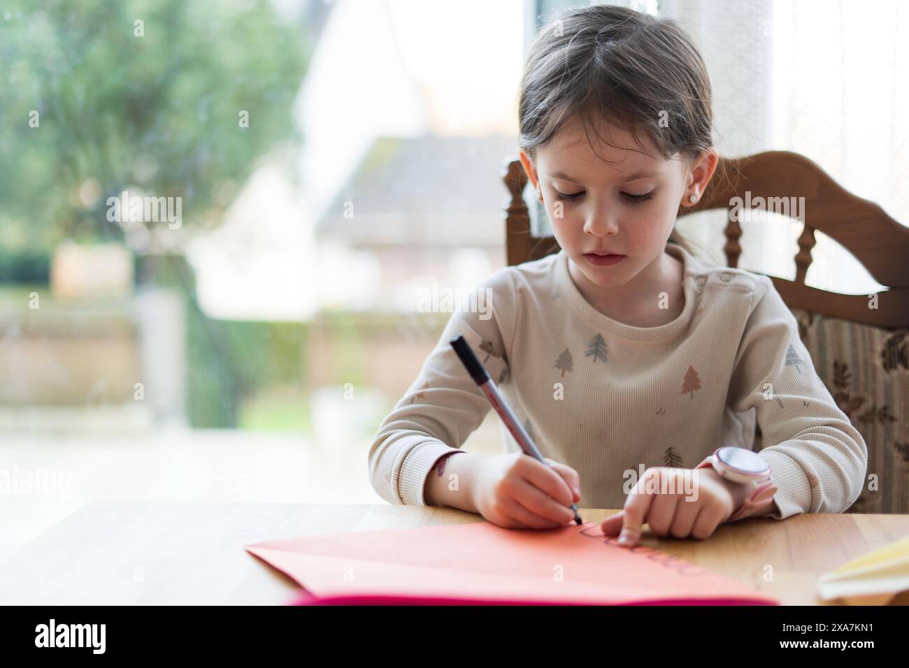 Une petite fille mignonne avec une queue de cheval, assise à une table de salon, fait ses devoirs, tenant un stylo dans sa main. Concept d'éducation à domicile. Banque D'Images