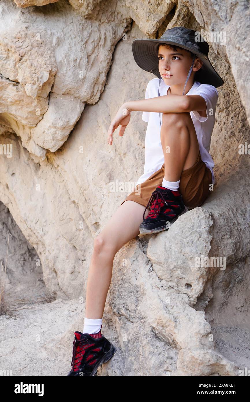 Garçon caucasien préadolescent avec chapeau et bottes de trekking sur le sentier de montagne assis sur un rocher reposant Banque D'Images