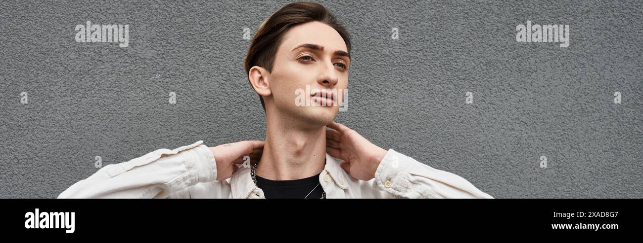 Un jeune queer prend une pose fière devant un mur gris, mettant en valeur sa tenue élégante et célébrant la fierté LGBT. Banque D'Images