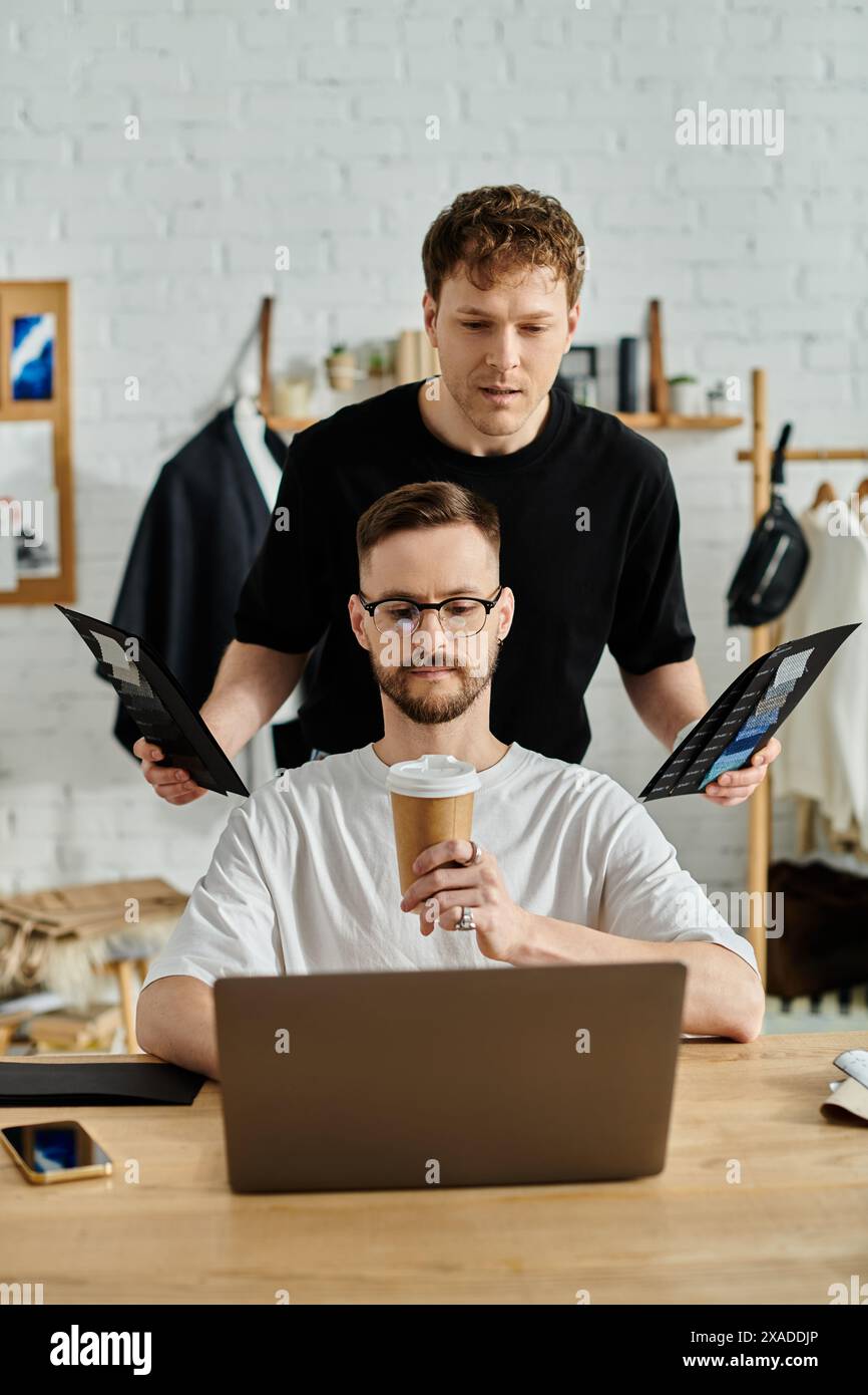 Un homme est assis à une table, engrossé dans son ordinateur portable, tout en travaillant sur la conception de vêtements tendance pour son atelier de mode. Banque D'Images