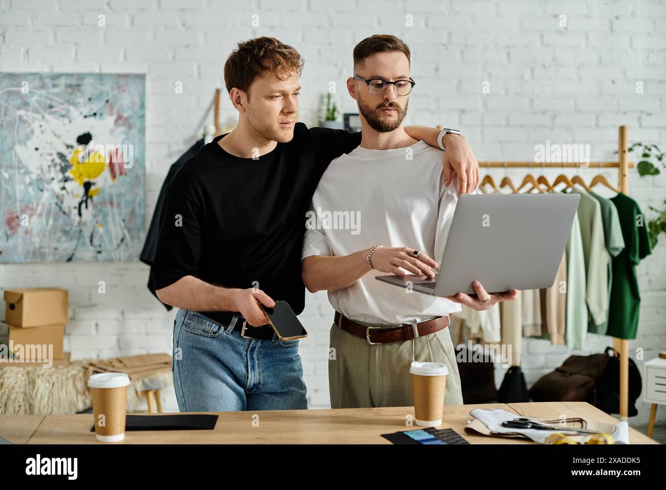 Un couple gay se tient côte à côte devant un ordinateur portable, travaillant passionnément ensemble sur des vêtements tendance dans un atelier de créateurs. Banque D'Images