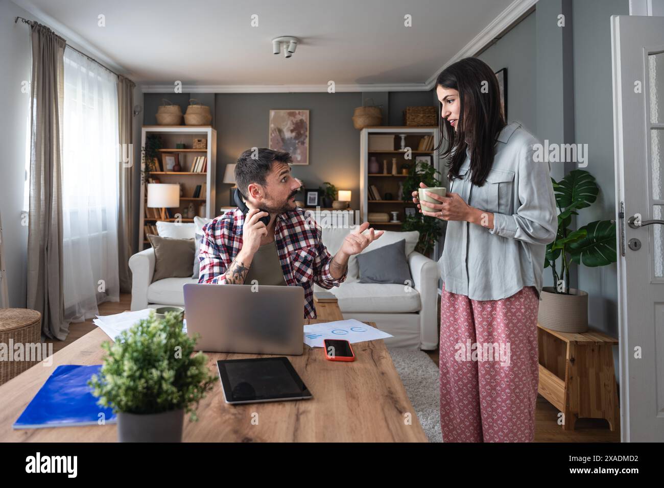 Jeune femme apportant tasse de café pour son petit ami alors qu'il travaille en ligne sur ordinateur portable à table à la maison. Homme d'affaires indépendant ayant un appel vidéo avec Banque D'Images