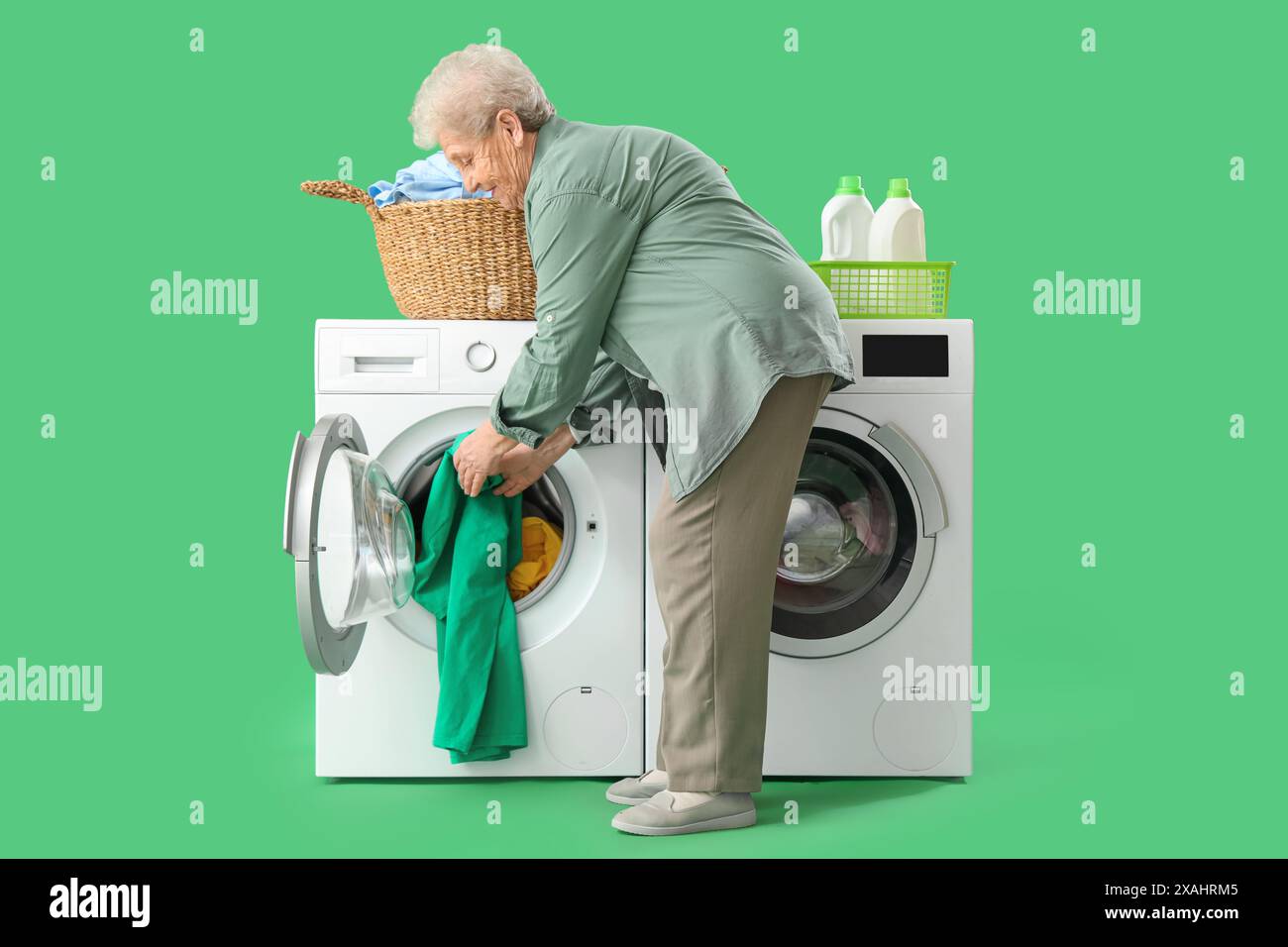 Femme senior avec panier de vêtements mettant le linge dans la machine à laver sur fond vert Banque D'Images