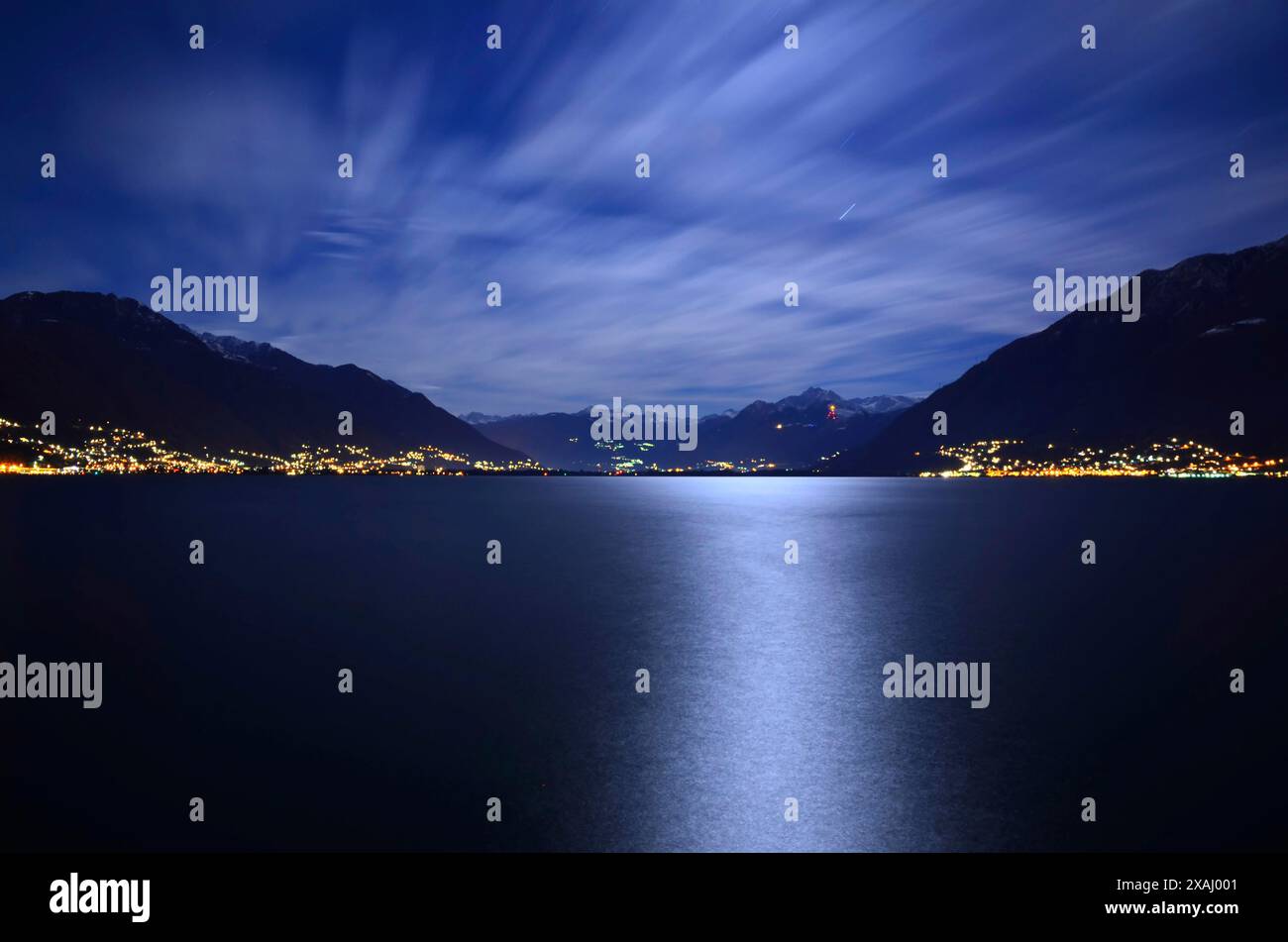 Belle Lune lumière au-dessus d'un lac alpin majeur avec montagne enneigée et nuages en longue exposition la nuit à Locarno, Tessin, Suisse Banque D'Images