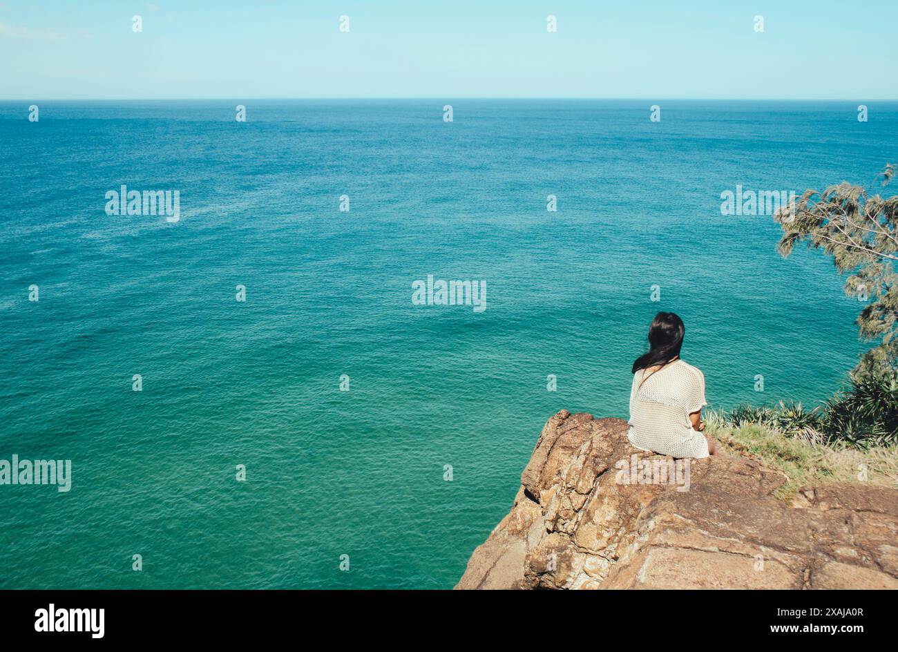 Jeune femme décontractée assise sur un rocher seul regardant vers la mer turquoise, fond de vacances d'été, voyageur solo de derrière appréciant la vue Banque D'Images