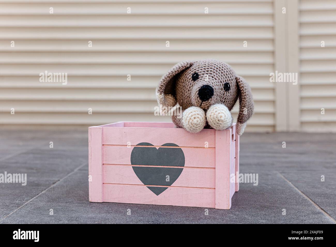 Chiot tricoté dans une boîte rose en bois avec un coeur dessus. Chien jouet crocheté mignon avec sa tête sur ses pattes regardant avec tristesse. Adoptez le concept de ne pas magasiner Banque D'Images
