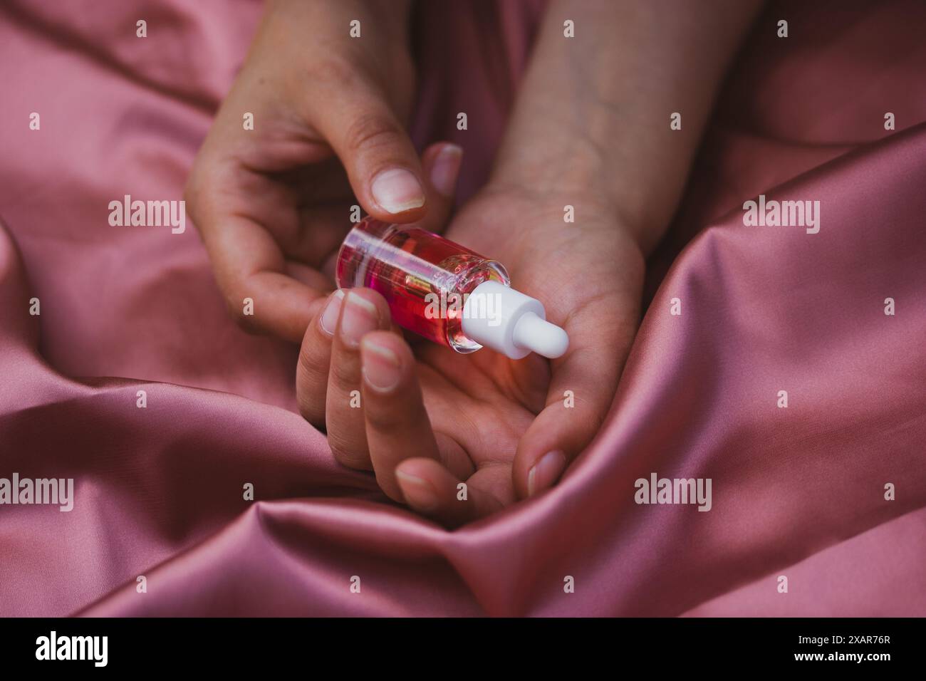 Flacon pipette en verre transparent avec sérum rouge pour le visage, soins du corps dans les mains sur textile rose soyeux. Soins de beauté féminins. Produit esthéticien Banque D'Images