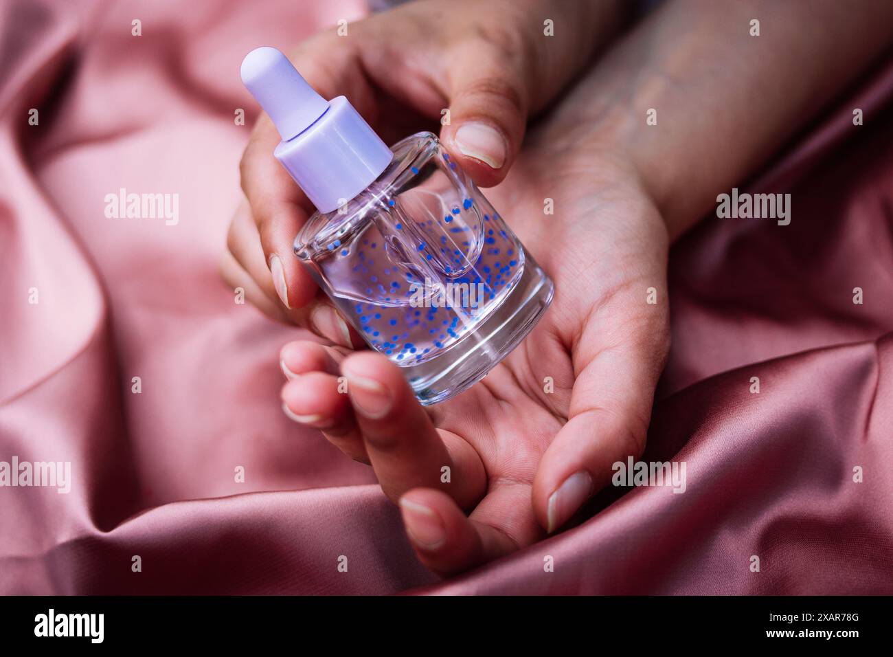 Flacon pipette en verre transparent avec sérum violet pour le visage, soins du corps dans les mains sur textile rose soyeux. Soins de beauté féminins. Produit esthéticien Banque D'Images