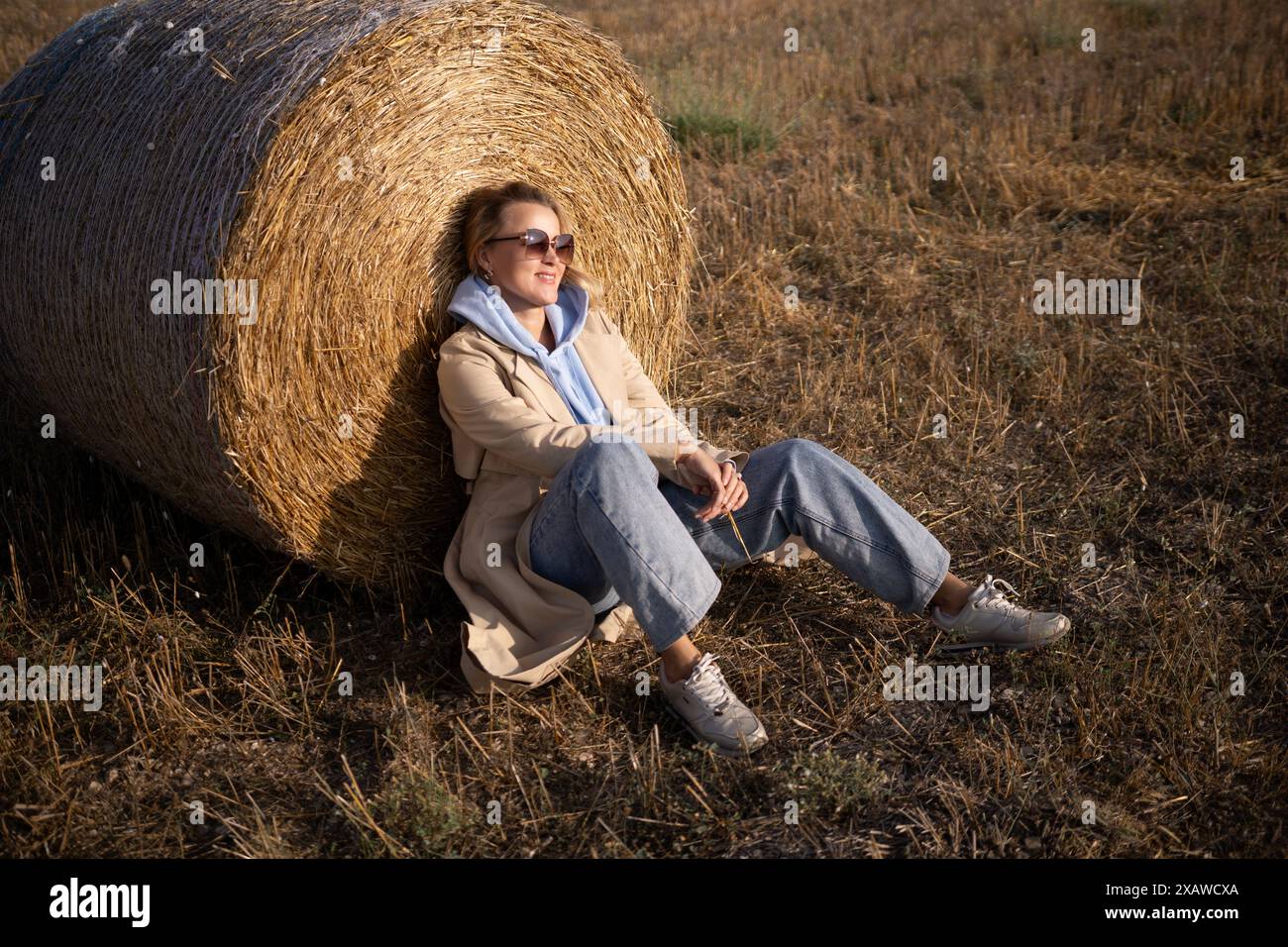 Une femme est assise sur une balle de foin dans un champ. Elle porte une veste bleue et un Jean bleu Banque D'Images