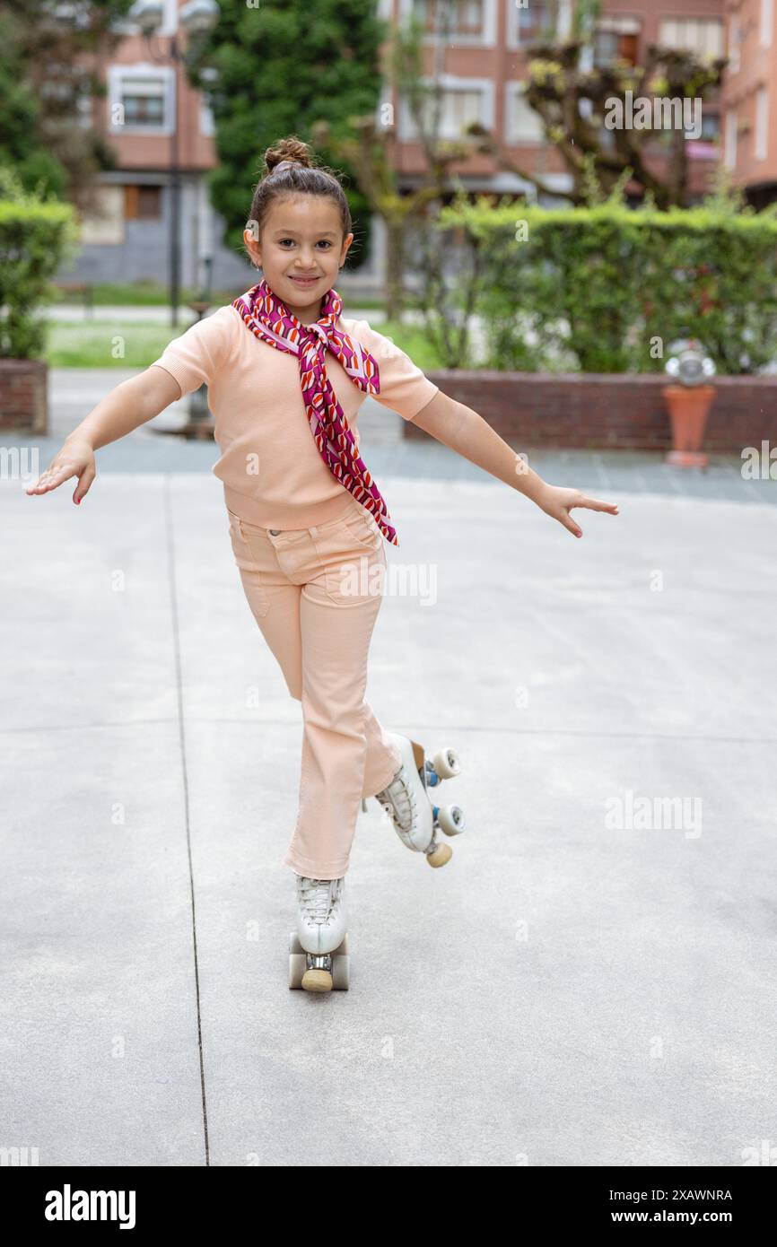 Une jeune fille joyeuse vêtue de vêtements couleur pêche et d'une écharpe à motifs sourit à la caméra tout en patinant à roulettes et en posant dans le parc. Banque D'Images