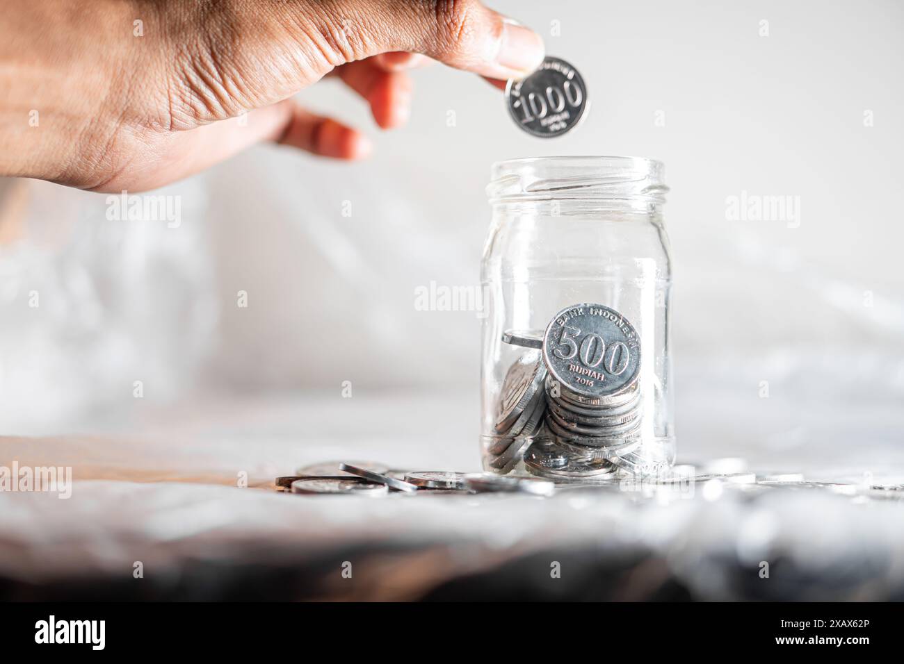 Une main plaçant une pièce de 500 Won dans un bocal en verre rempli d'autres pièces de 500 Won. Le pot est sur une surface blanche avec plus de pièces dispersées autour de lui. Banque D'Images