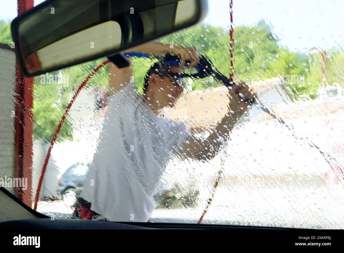 Vue depuis le siège passager d'un homme nettoyant le capot et le pare-brise d'une voiture à l'aide d'un pistolet nettoyeur haute pression. Banque D'Images