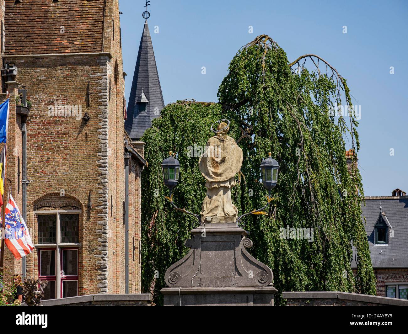 Statue en pierre devant des bâtiments historiques et une tour d'église, encadrée par de vieux arbres, sous un ciel bleu clair, Bruges, Belgique Banque D'Images