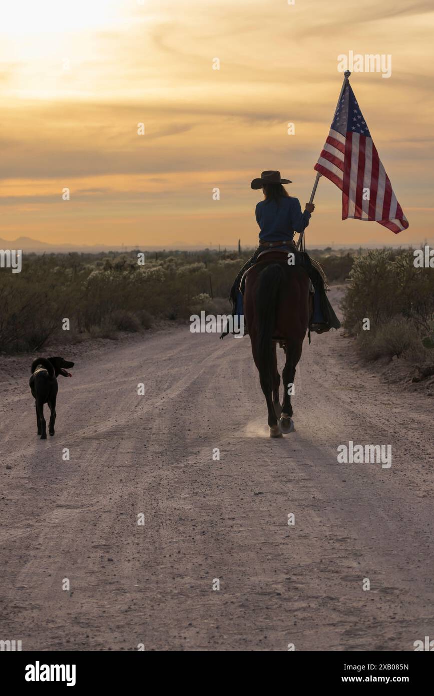 Main de ranch femelle, ou cowgirl, à cheval et portant le drapeau américain dans le coucher du soleil sur la route de terre sur un ranch en Arizona, États-Unis. Accompagné d'un chien. Banque D'Images